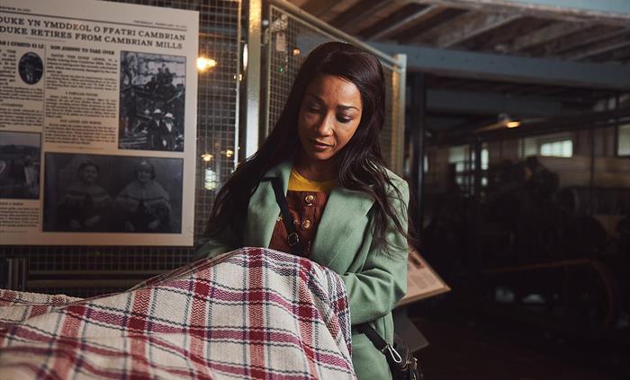 A lady wearing a green coat is lifting up a section of woollen cloth. It is finely woven in a red and cream check pattern, traditional to Cambrian Mil